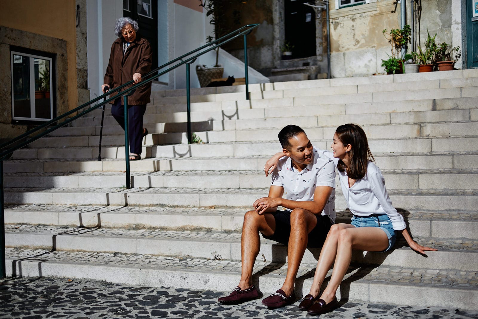 an elderly woman looks at Eliza Sam and Joshua Ngo as they're sitting on some stairs in a traditional Lisbon neighbourhood as part of a photoshoot with Lisbon Photographers Your Story in Photos