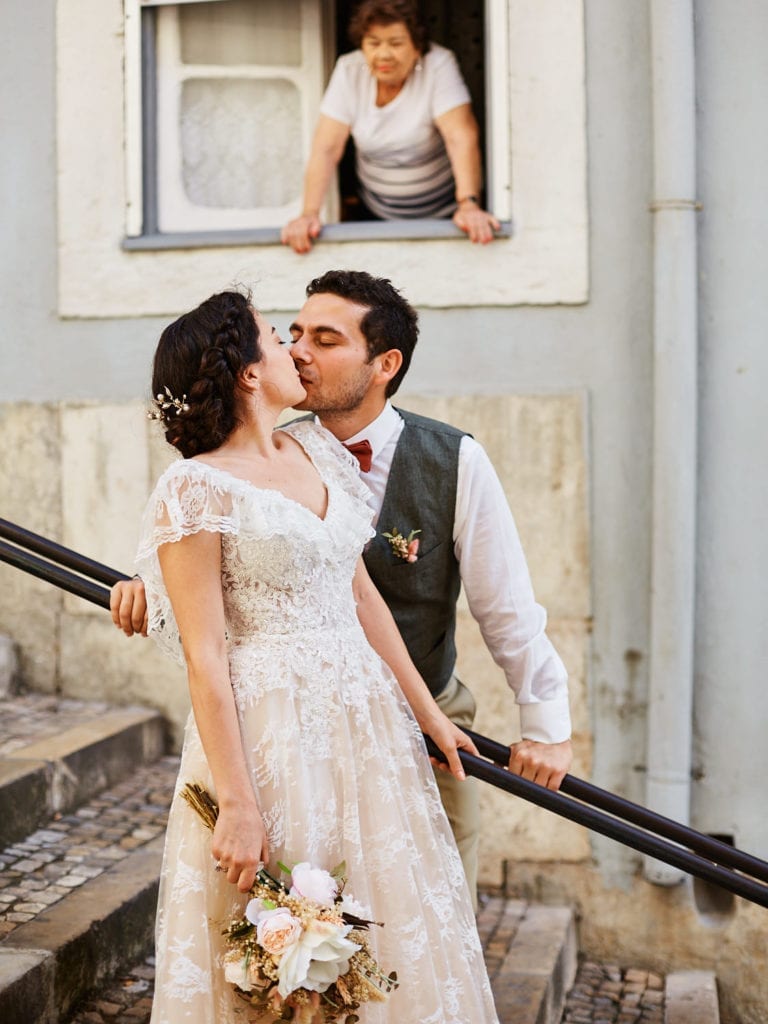 A couple in their wedding dress kiss on a stairway in the Mouraria neighbourhood in Lisbon, Portugal, while an elderly lady looks at them from the window | Photo taken during a Lisbon Elopement by Lisbon Photographer Your Story in Photos