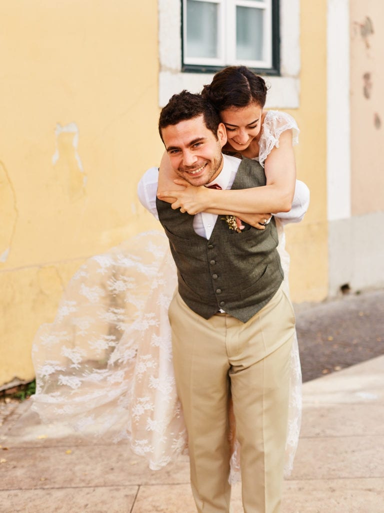 a groom gives his bride a piggyback ride in the streets of Alfama, in Lisbon, Portugal, on their elopement